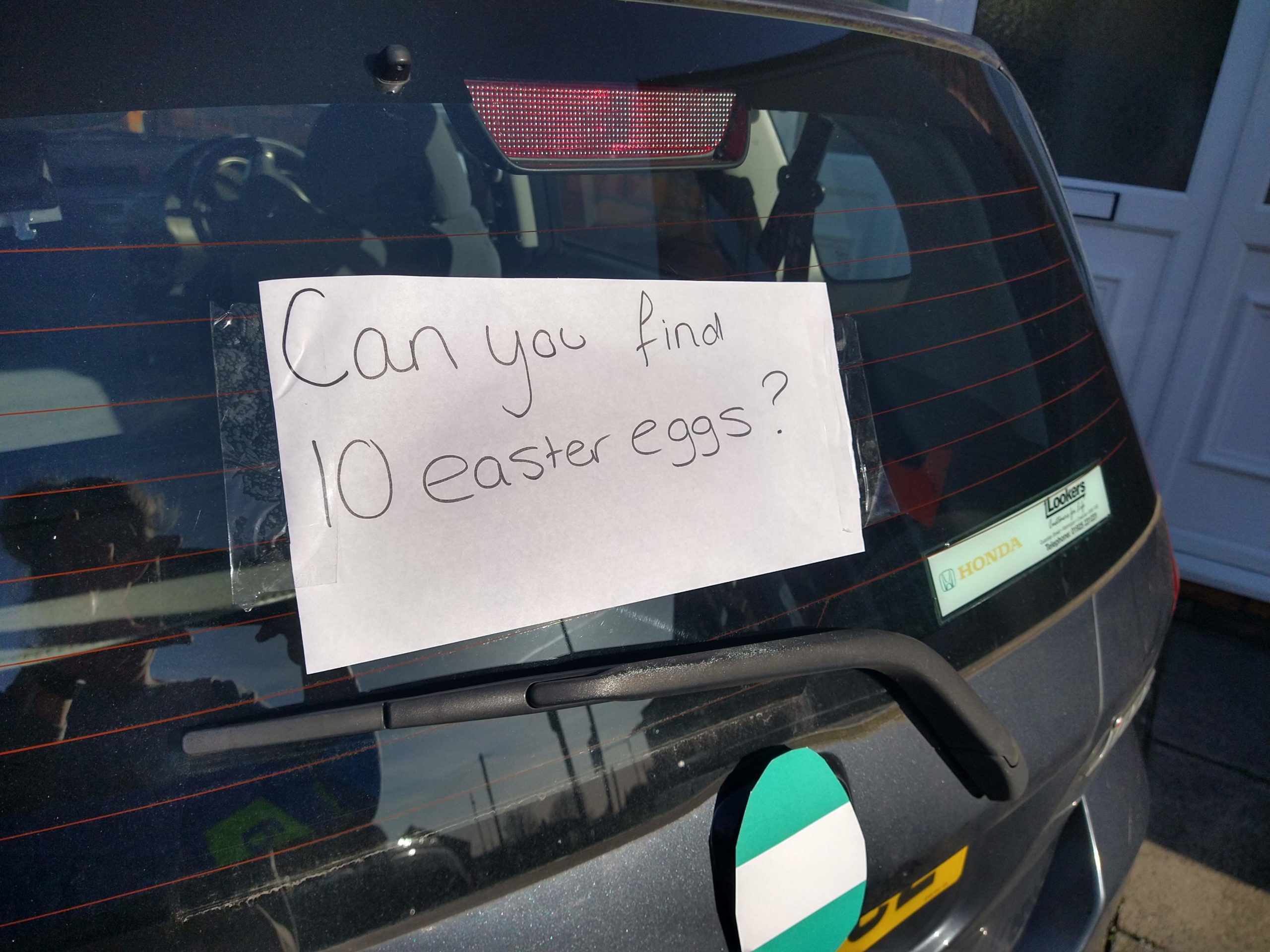 A car window with a aper sign in the window saying 'Can you find 10 easter eggs?' there is a turquoise peice of caedboard shaped like a large egg with a white stripe.