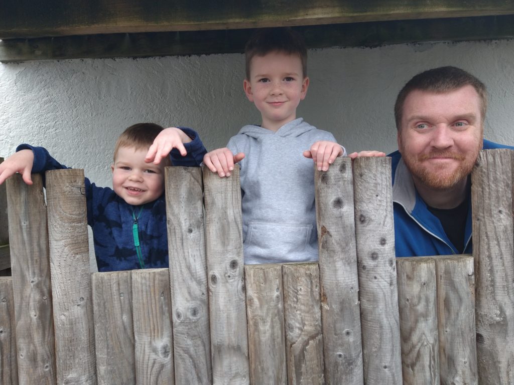 A toddler, young boy and dad with a beard smile in a wooden fort.