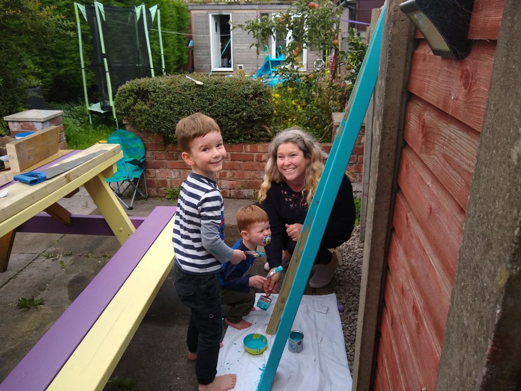 A mum, a boy and a toddler are painting a plank of wood blue next to a multicoloured bench