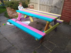A brightly multi coloured bench in a garden a boy stands proud next to it