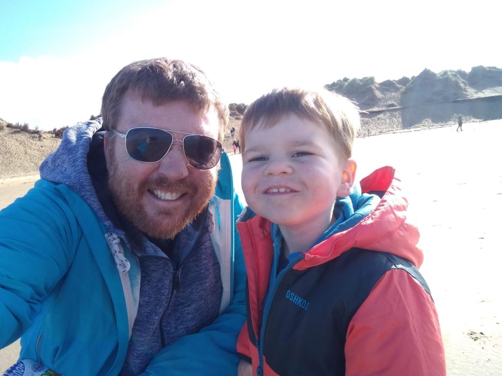 A dad with a beard and sunglasses with a young boy sit smiling on the beach