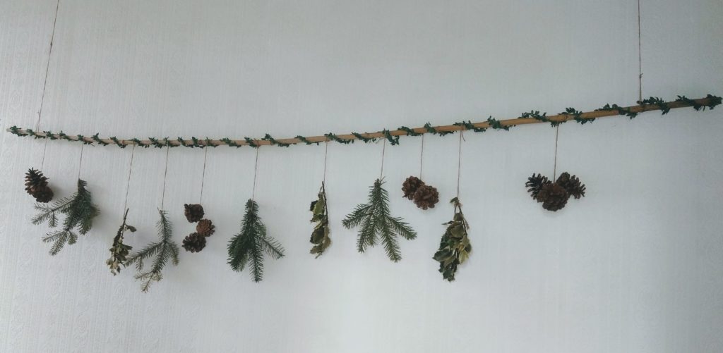 A bamboo cane wrapped in leaf wine hung in a hallway. Hanging from it with twine are pine branches, pine cones and holly twigs.