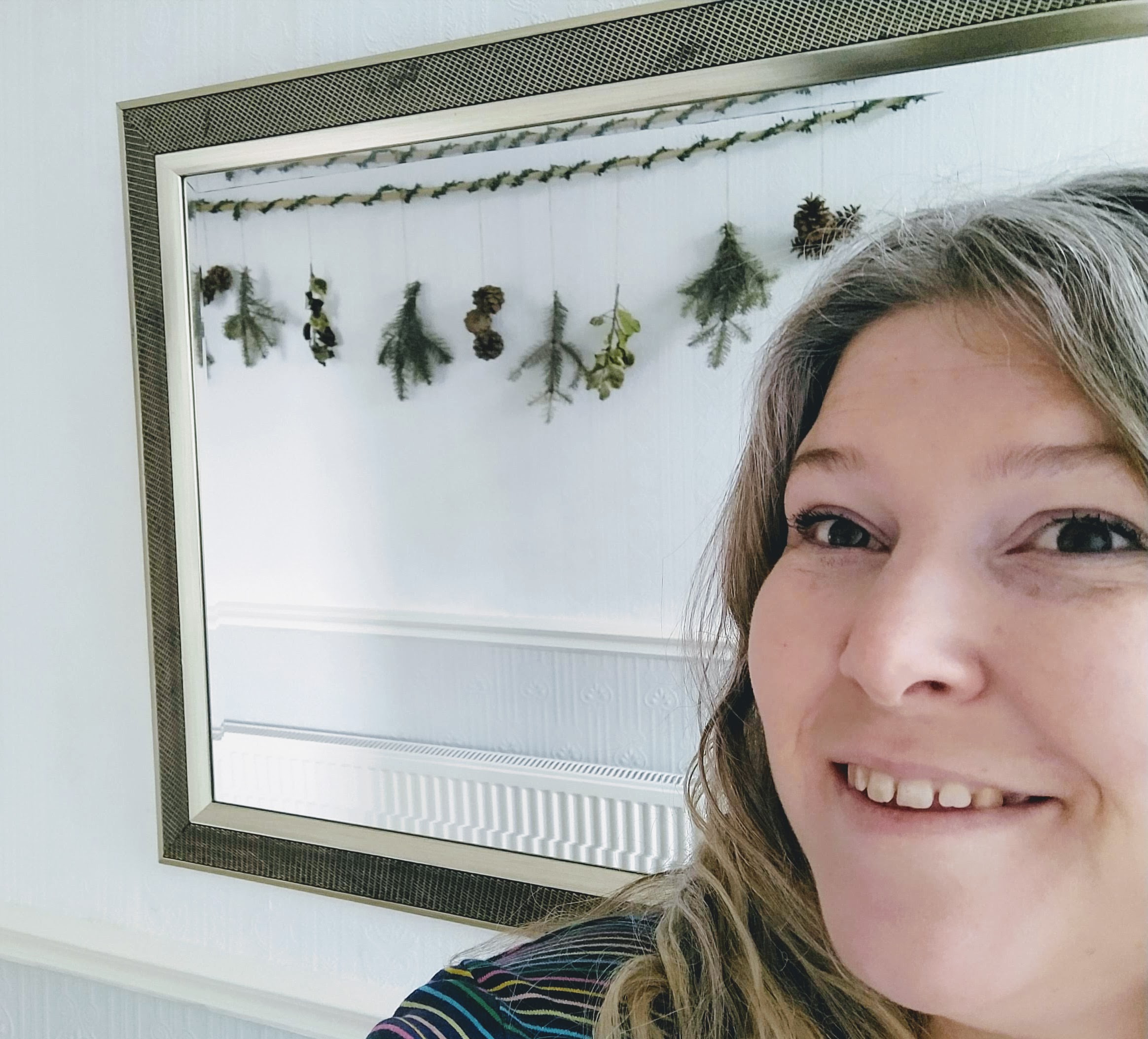 A bamboo cane wrapped in leaf wine hung in a hallway. Hanging from it with twine are pine branches, pine cones and holly twigs. This is reflected in a bronzed framed mirror with a middle aged women smiling in the frame. She looks tired and is unphotogenic but proud