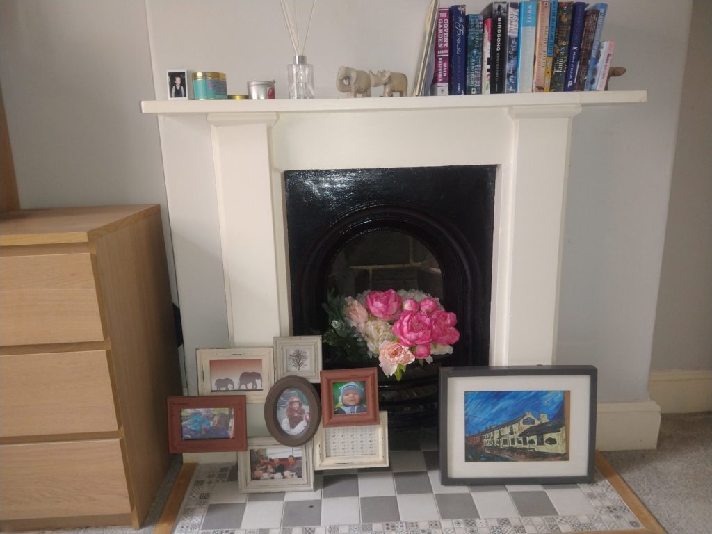A fireplace with a white surround and black grate. Inside the grate is a bunch of artificial flower with dark and light pink. In front of the fire place are photo frames. One of the family members and the other is a picture of a pub hand painted. The mantle piece is cluttered and has photos, candles, a fragrant infuser, statues of an elephant, rhino next to a stack of books with a statue of a hippo hiding at the end. 