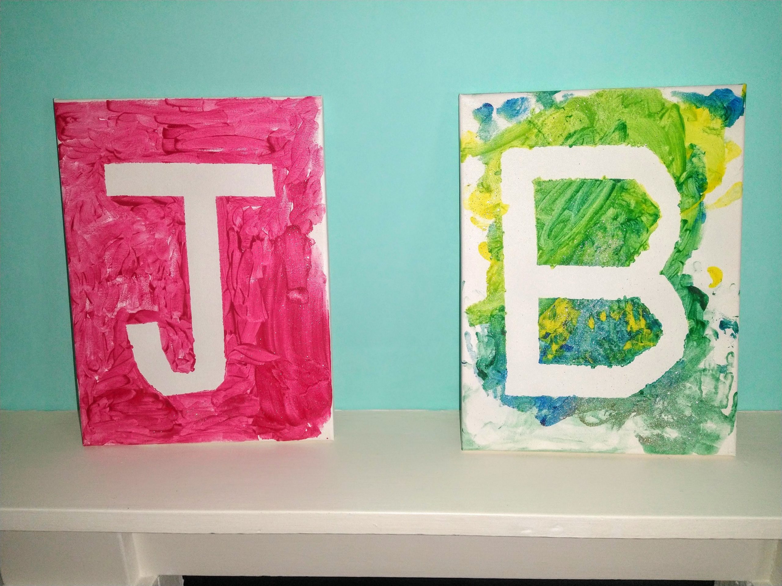 Two large canvas boards sit on a white mantle piece against a turquoise wall. They are painted with the initials on. The first is painted red with a J and has been painted with accurancy and small strokes. The second is the letter B. The background is painted with multiple colours blue, green and yellow with glitter over the top. This has been painted haphazardly but looks wonderful.