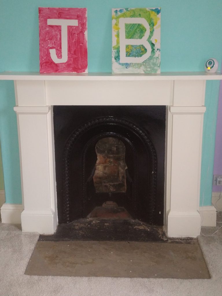 A white fireplace surround with a black interior that is empty. The floor before the fireplace is bare. The mantle piece had two canvas boards that have been painted to show initials J and B. One board is red and the other multiple colours -yellow,green and blue with silver glitter.