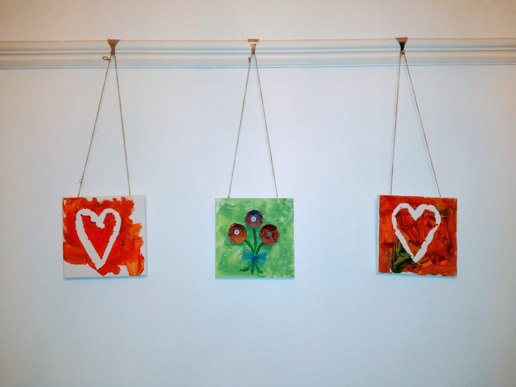 A white wall with three small canvas boards hung up using twine from a picture hanging rail with hooks. Children have painted these. One is a picture of three flowers using egg cartons and buttons and the other two are white hearts with a red background and red middle. The pictures are imperfect but beautiful