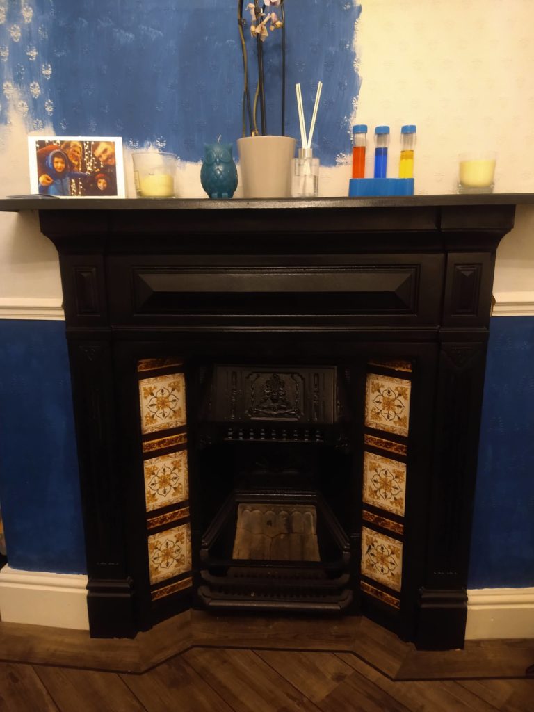 A black fireplace with an empty black grate. There are half painted blue walls and only half done. But i cannot be bothered as it was lock down and I just watched tv instead. On top of the mantle piece are pictures and items from a science kit like test tubs.