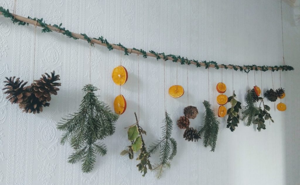 A hanging feature in a hallway. A garden cane wrapped in leaf wire. Hanging with twine are pine cones, pine branches, dried orange slices and holly branches and leaves.