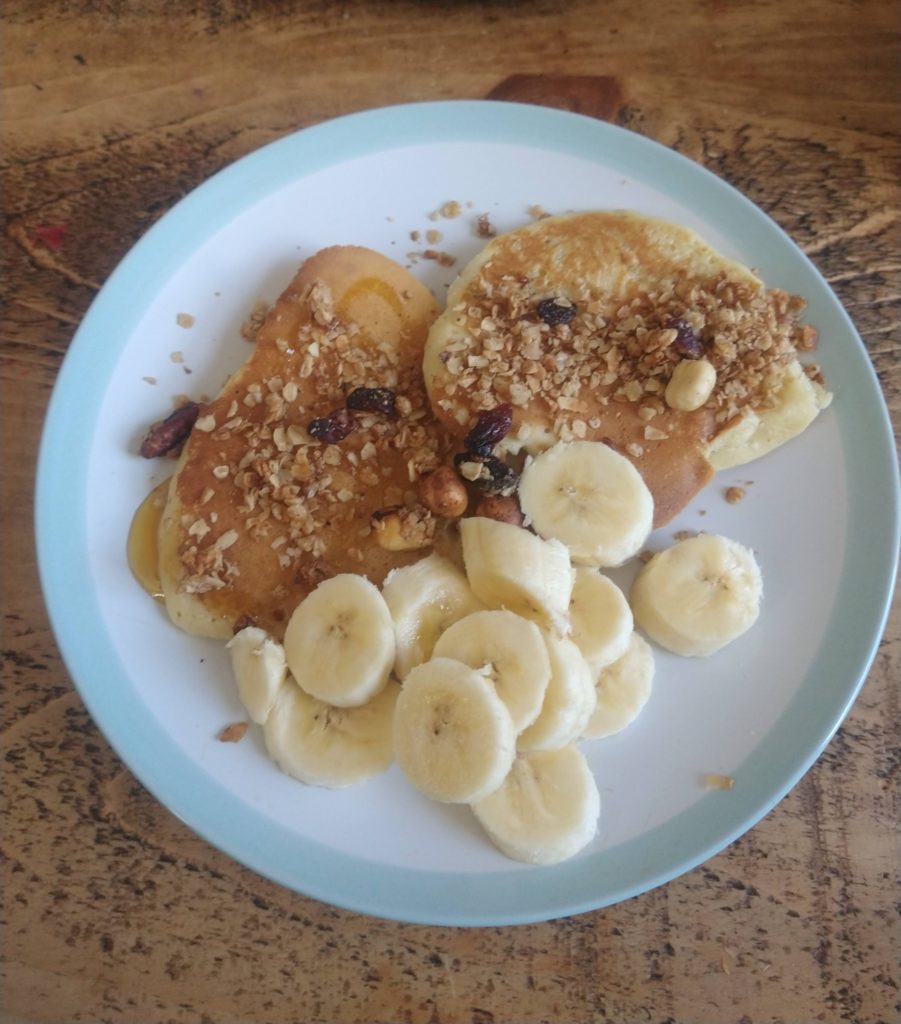 Two american pancakes on a white plate. Next to a sliced banana and topped with golden syrup and homemade granola.