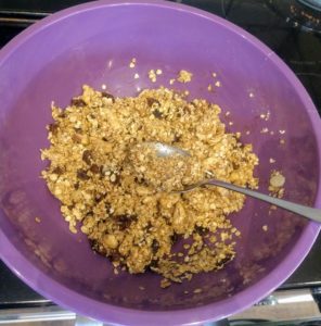 A purple bowl with the granola ingredients mixed together with a tablespoon in the mix.