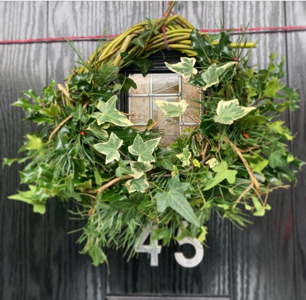 A beautiful fresh green wreath. Made of vines and ivy hung on a black front door with the number 45 .