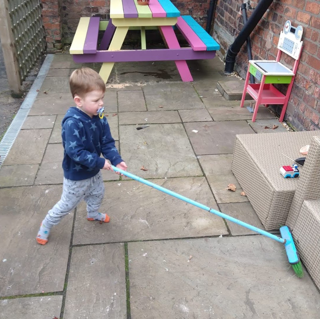 A young toddler with a dummy is sweeping outside with a long handled blue brush.