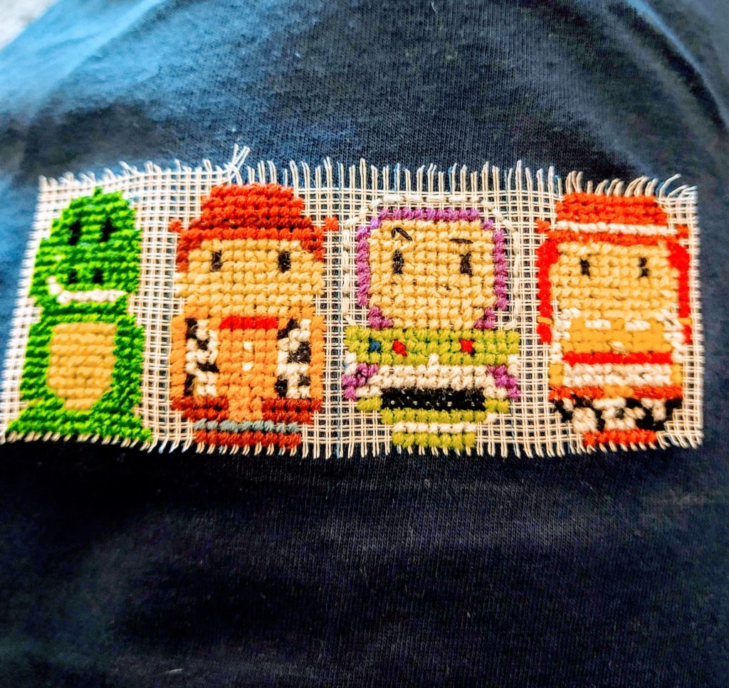 A half finished cross stitch pattern on a tshirt. It is a picture of characters from toy story. From left to right - rex, woody, buzz light year and Jessie.