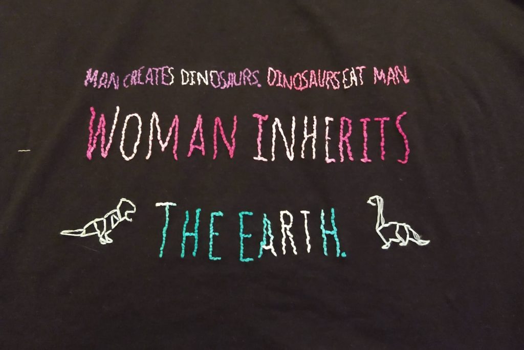 A black tshirt with ombre lettering read man creates dinosaurs, b=dinosaurs eat man, women inherit the earth. with two small dinosaurs. 