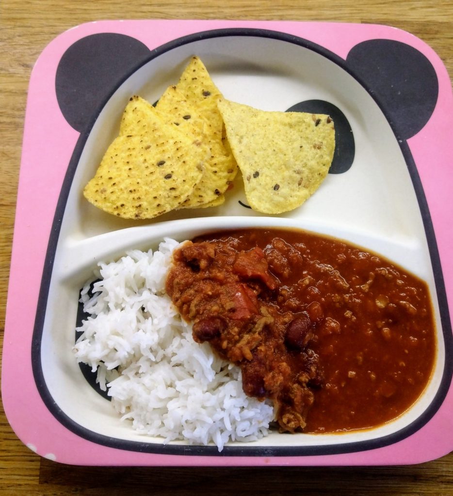 A bamboo panda plate with a chilli made with red lentils and mince with a side of rice and some tortilla chips on the side 