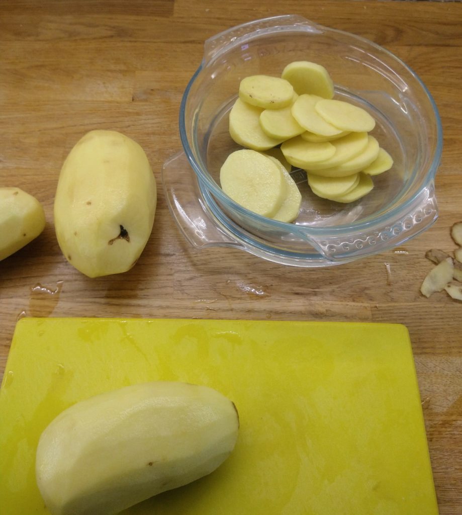 A peeled potato sits on a yellow chopping board above it another peeled postato sits on a wooden work top and a sliced peeeled potato sits in a circulare pyrex bowl 