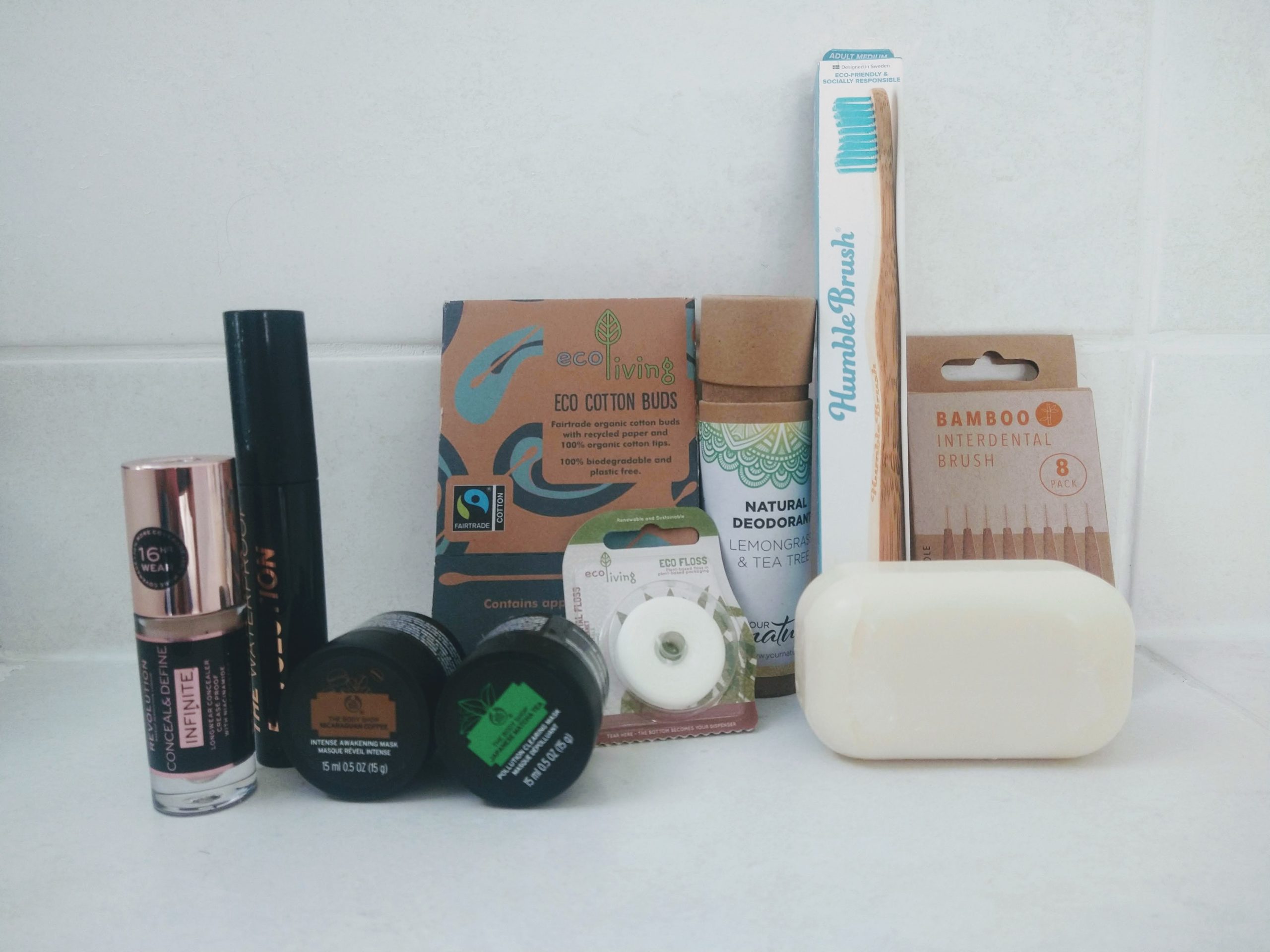 A white tiled background shows sustainable alternative beauty and health care products. Revolution make up, body shop face masks, plastic free cotton buds and dental floss, a bamboo toothbrush and a bar of soap.