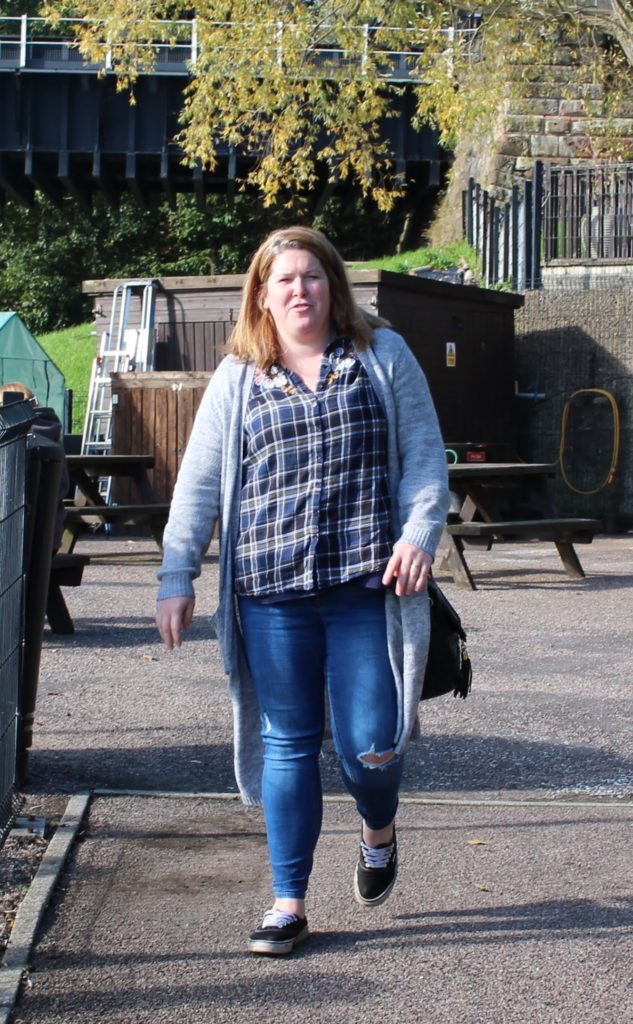 An unflattering piture of a mum in a checked shirt and cardigan walking through a park.