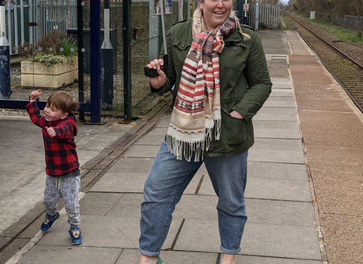A size 18 mum holding a litter oicker in a green linen jacket stood at a train station.Stood next to her is her 3 year old son in a red and black checked jumper