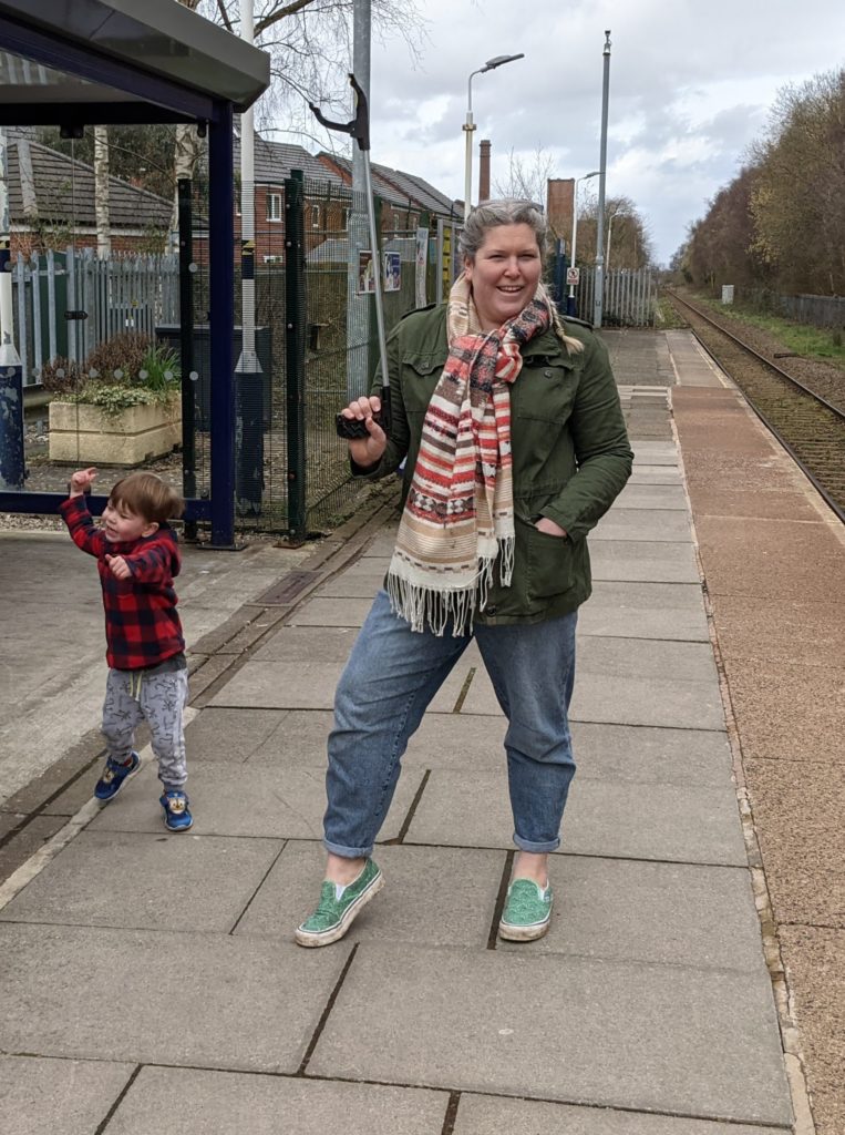 A size 18 mum holding a litter oicker in a green linen jacket stood at a train station.Stood next to her is her 3 year old son in a red and black checked jumper