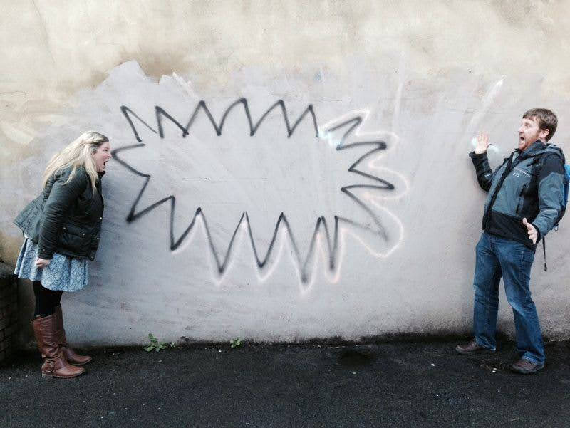A lady in a skirt looking like she is screaming at a man who looks shocked. Behin=d them on a wall is a jaggered shape spray painted on the wall that looks like an angry speech bubble