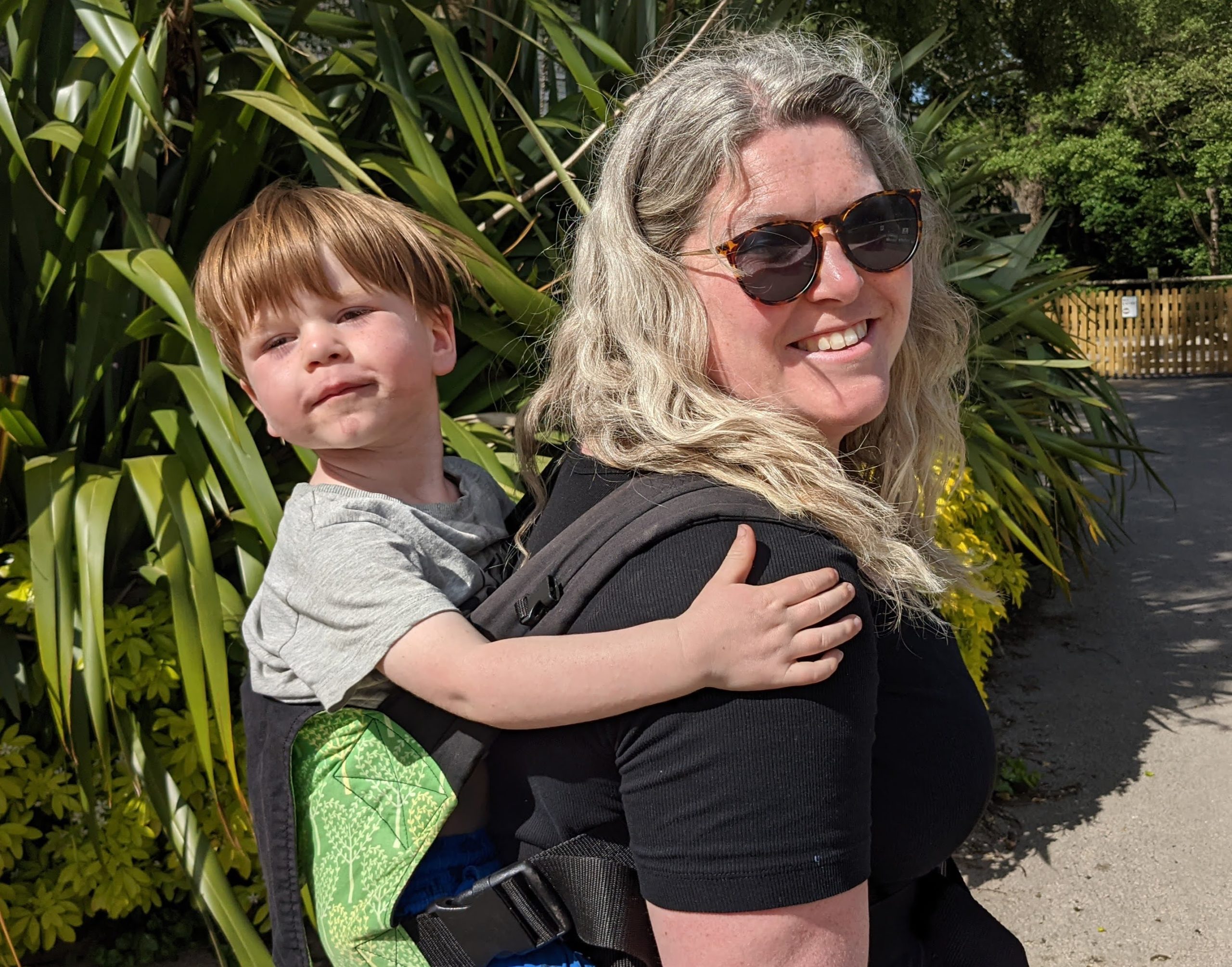 A 34 year old mum with grey hair, wearing sunglasses and smiling. On her back she caries her son in a green sling. They are outside at Drayton Manor and are enjoying their day. She is happy.