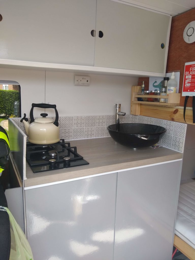 A view of the small kitchen area.  A two ring gas hob with a cream kettle on top. and a black glass bowl sink sits ontop of the counter wih a large tap.there are cupboards above 