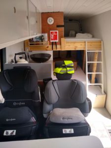 Looking down the now converted van. There are two seats with car seats for child in. A view of a small kitchen and he two bunk beds and ladder