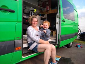 A mum and son sit in the side door way of a large green van. They are smiling and happy.