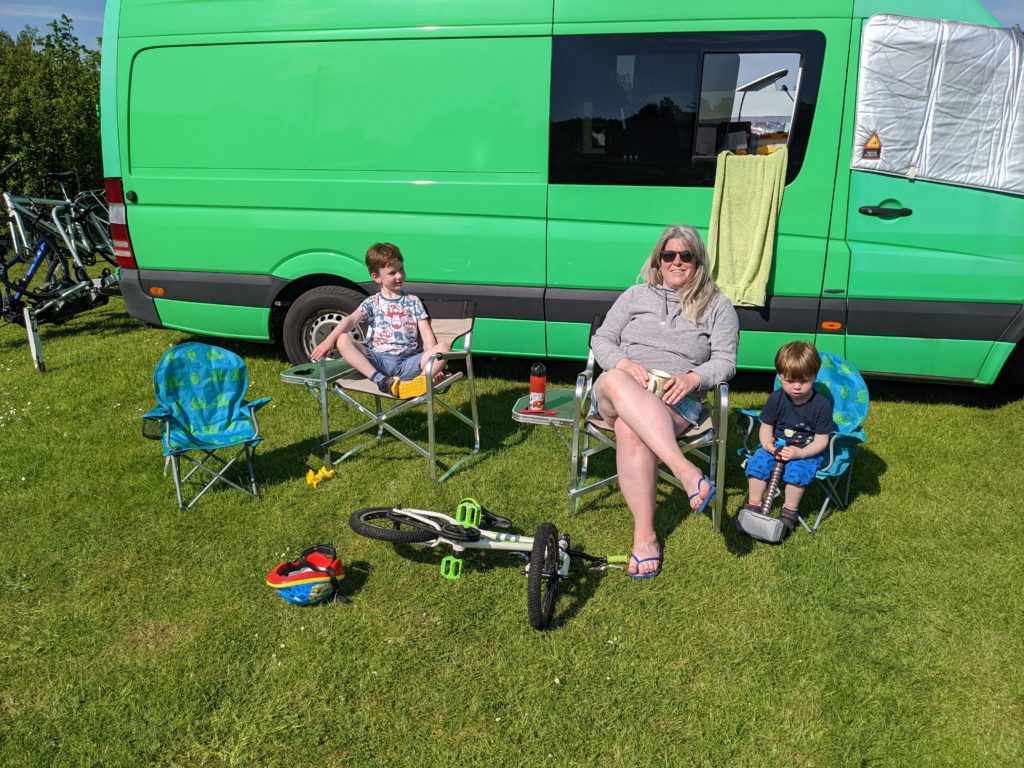 A family sit outside a green van on camp chairs smiling in the sunshine.